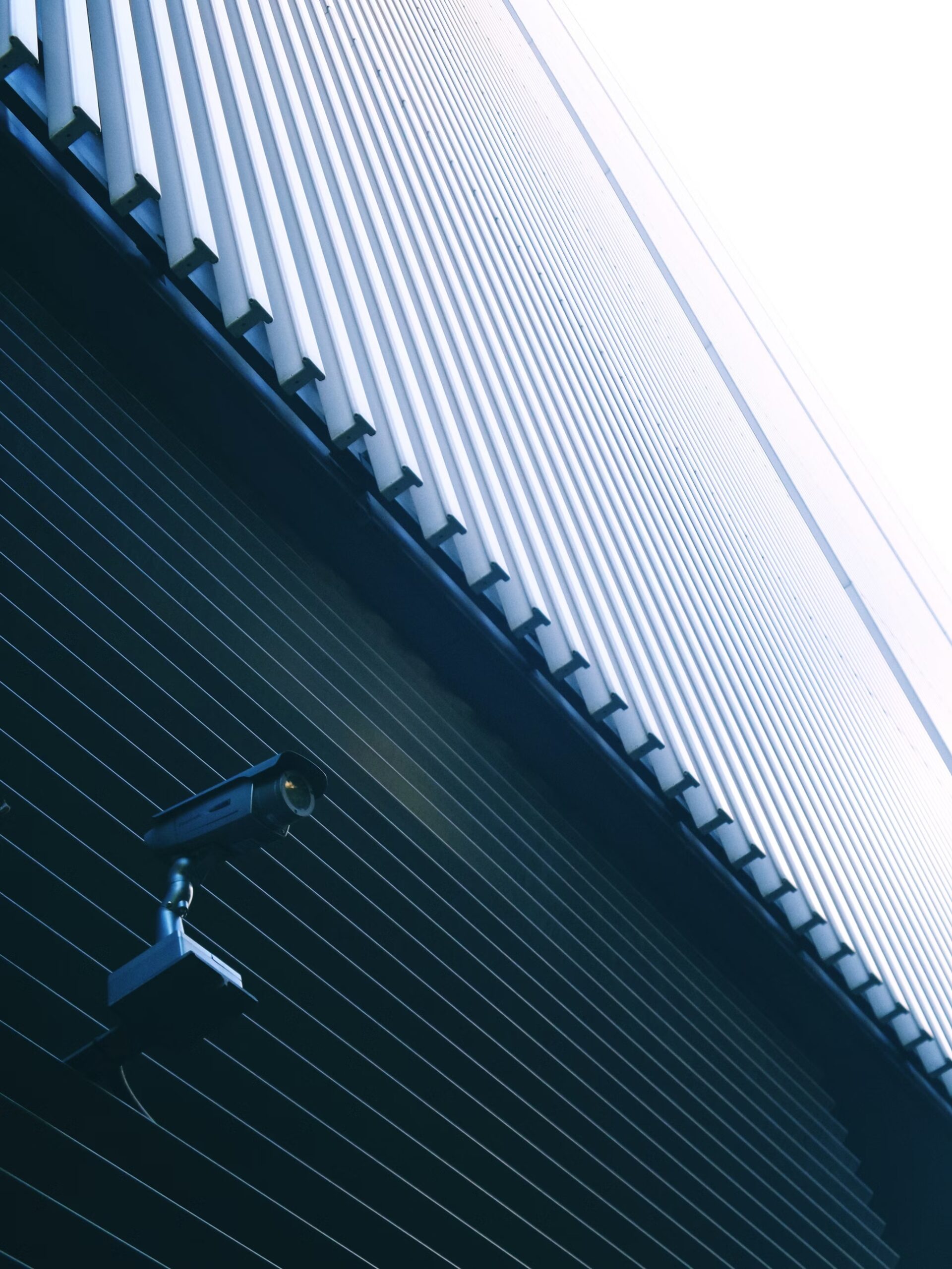 Una telecamera di sicurezza montata su una parete scura sotto un edificio alto e moderno con pannelli metallici verticali, vista da un'angolazione bassa contro un cielo luminoso.